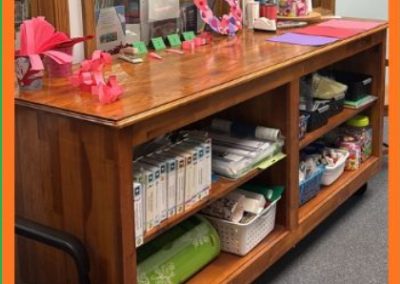 A wooden desk with crafts on top and organized supplies on shelves below, in front of windows showing books.